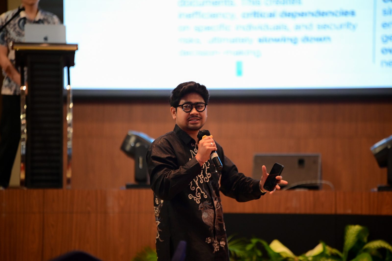 Laugiwa in a black batik shirt speaking into a microphone in front of a slide.
